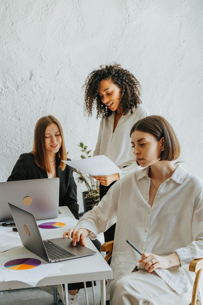 team-01 Three diverse businesswomen collaborating around laptops and documents in a modern office setting.